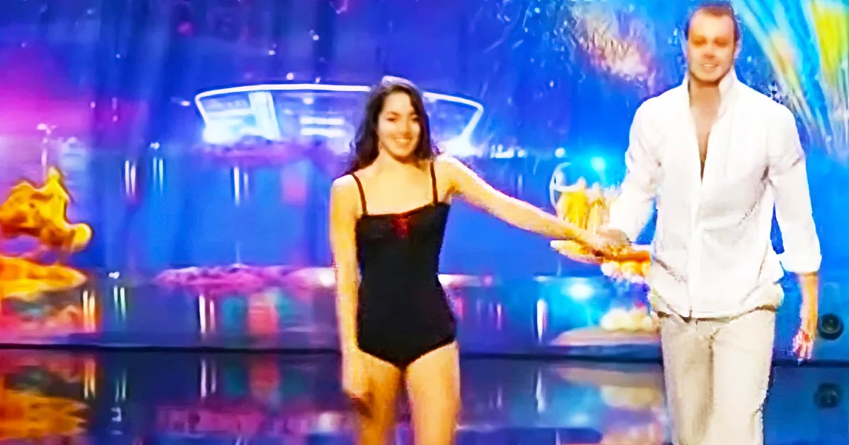 A woman in a black swimsuit is walking with a man in a white shirt on a stage with colorful lights.