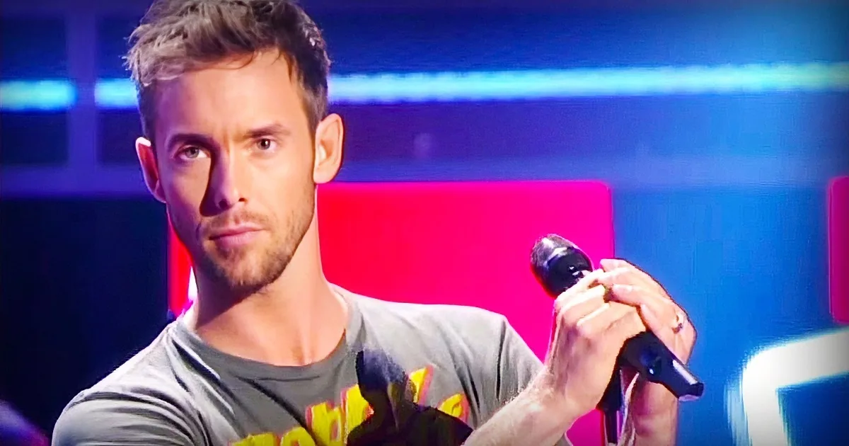 A man with short hair and a beard holds a microphone, wearing a gray t-shirt with yellow text, against a colorful background.