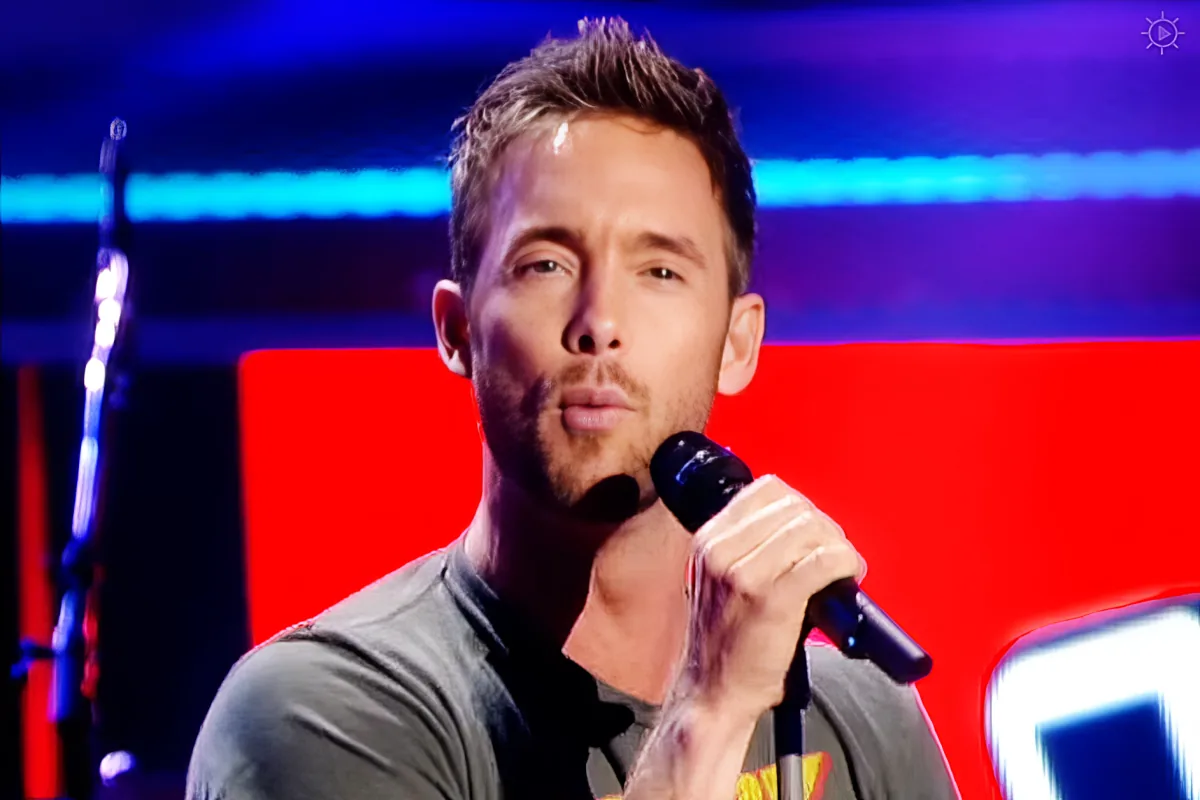 A man with short hair and a beard, wearing a gray shirt, is singing into a microphone on stage with a red background.