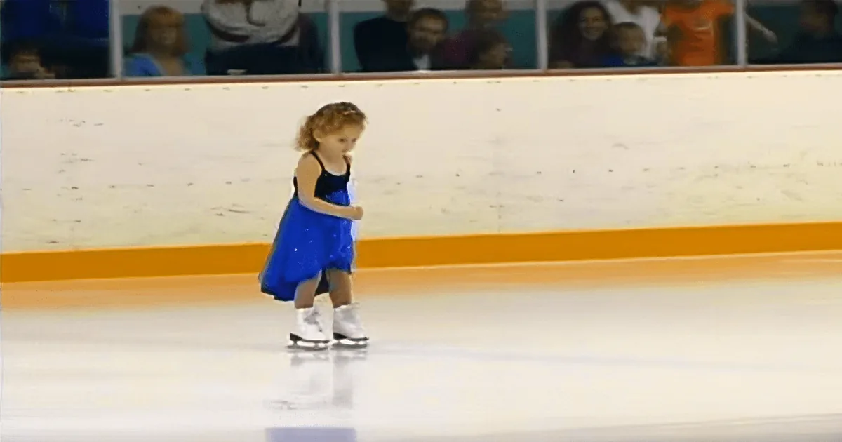 A young girl in a blue dress skates on an ice rink, surrounded by spectators.