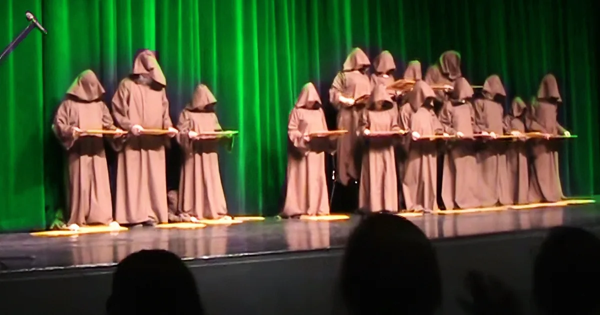 A group of people in brown robes stand in a line on a stage, holding objects, against a green curtain backdrop.