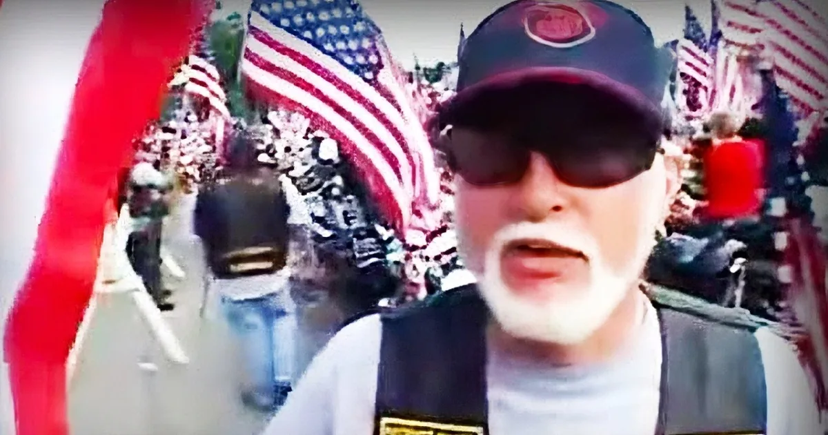 A man wearing a hat, sunglasses, and a white mustache stands in front of a crowd holding American flags.