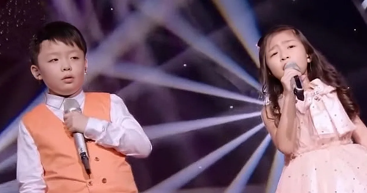 A young boy and girl are singing on stage, each holding a microphone, with a starry backdrop.