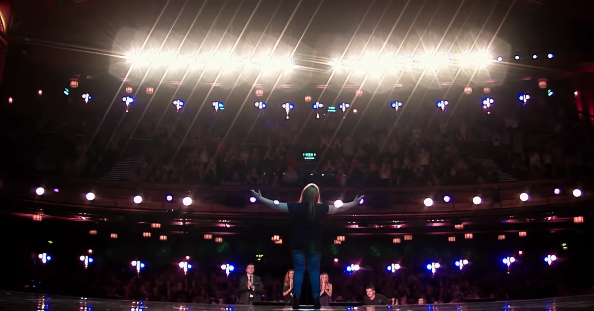 A person stands on stage with arms outstretched, facing a large audience illuminated by bright stage lights.