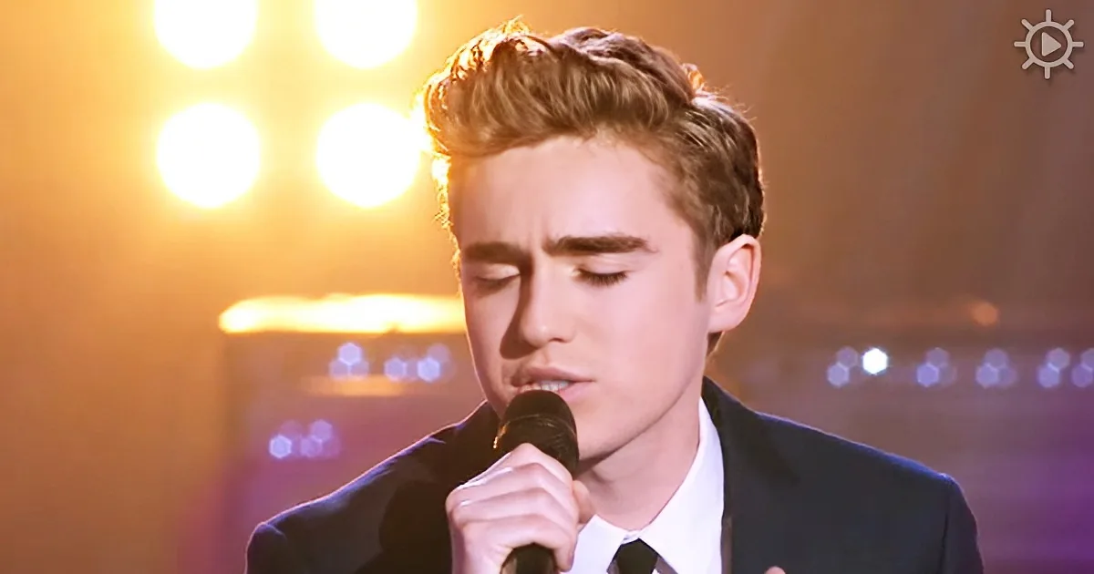 A young man in a suit sings into a microphone, eyes closed, under bright stage lights.
