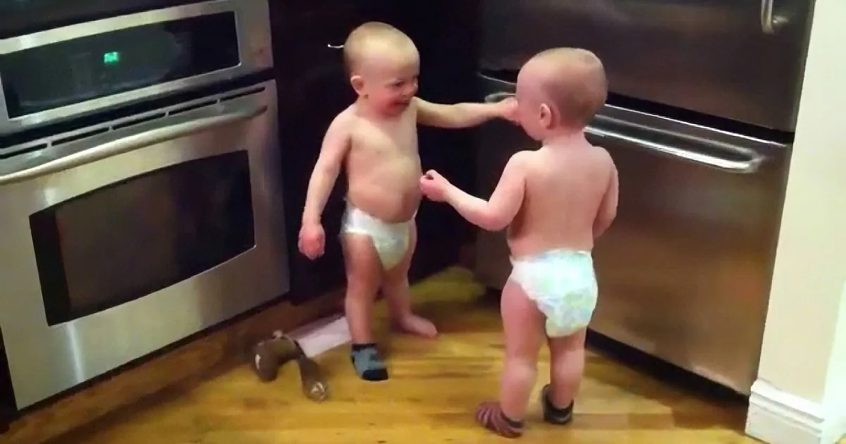 Two toddlers in diapers stand in a kitchen, one holding the other's arm, both looking at each other.
