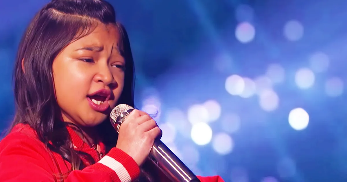 A person in a red outfit sings into a microphone against a blue, bokeh-lit background.