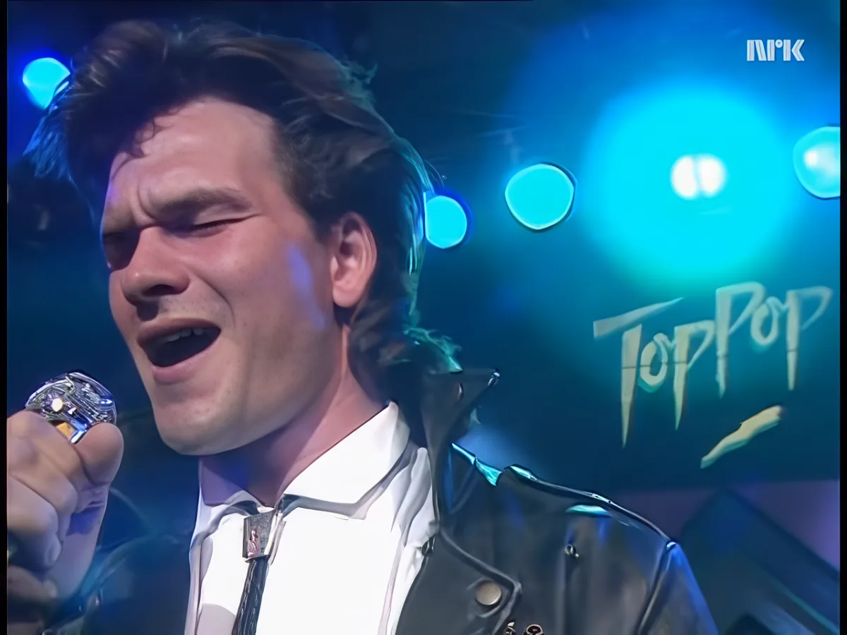 A man in a leather jacket and white shirt sings passionately on stage with blue lights and a microphone.