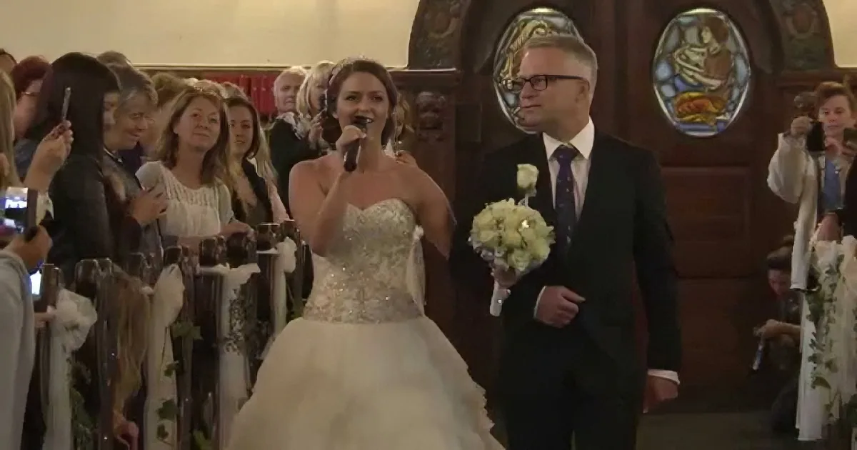 A bride in a white gown sings into a microphone as she walks down the aisle with her father.