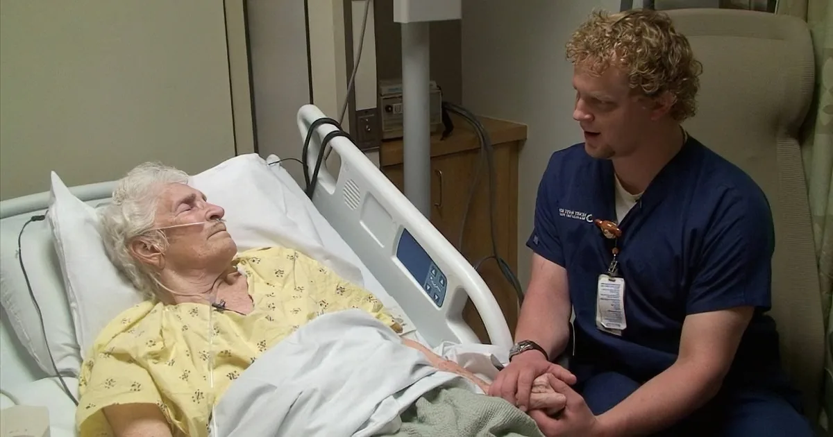 A nurse in scrubs holds hands with an elderly patient in a hospital bed.