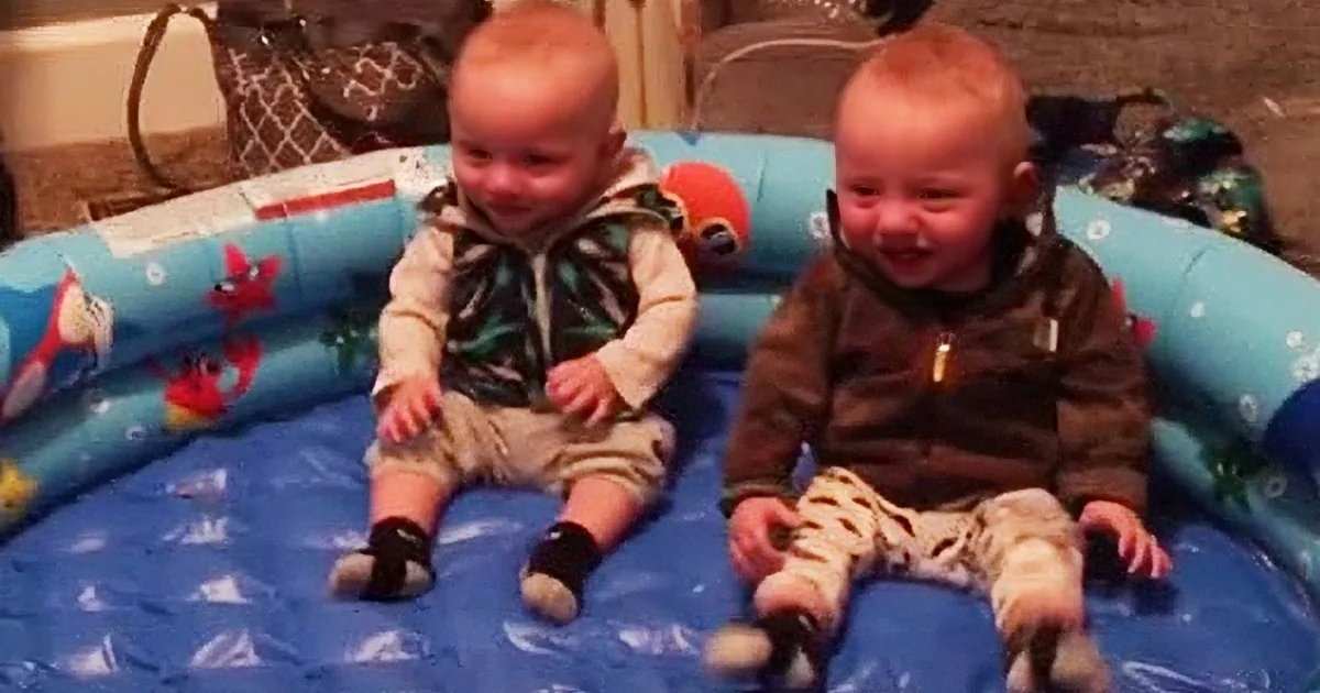 Two identical twin babies are lying in a blue inflatable pool, smiling and looking at the camera.