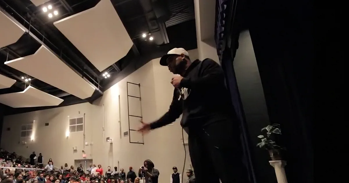 A man in a black suit and cap stands on a stage, gesturing to an audience in a large indoor venue.