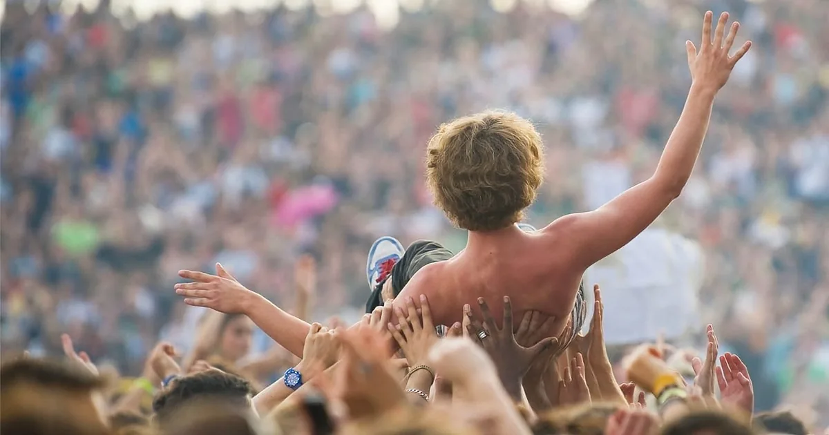 A shirtless person is crowd surfing, surrounded by raised hands and a large crowd in the background.