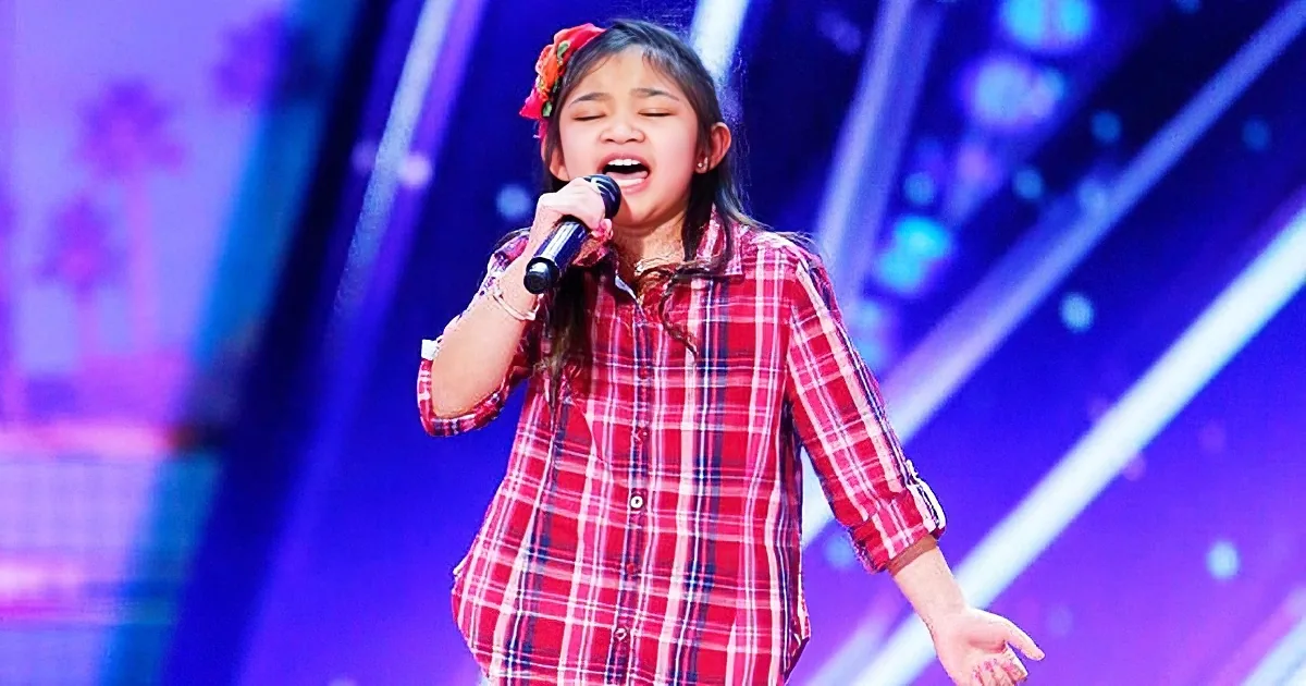 A girl in a red plaid shirt sings passionately on stage with a microphone, surrounded by vibrant purple and blue lights.