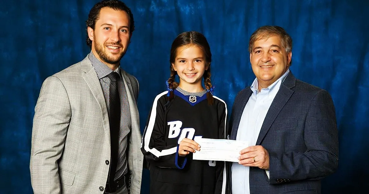 A young girl in a hockey jersey stands between two men in suits, one holding a check.