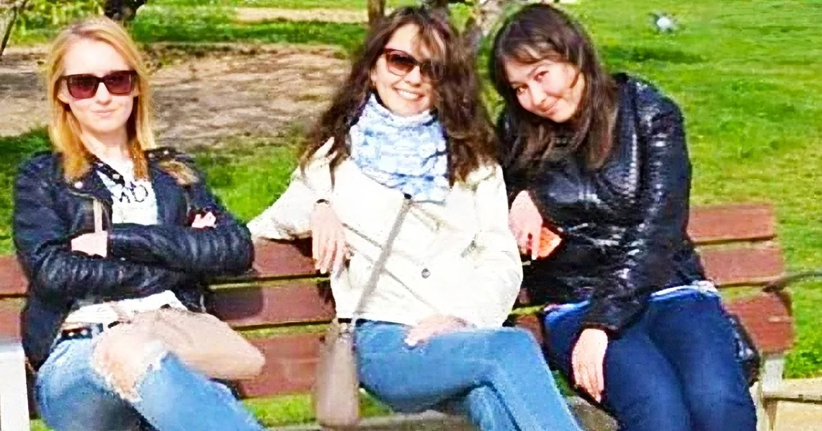 Three women in casual attire sit on a bench in a park, enjoying a sunny day.