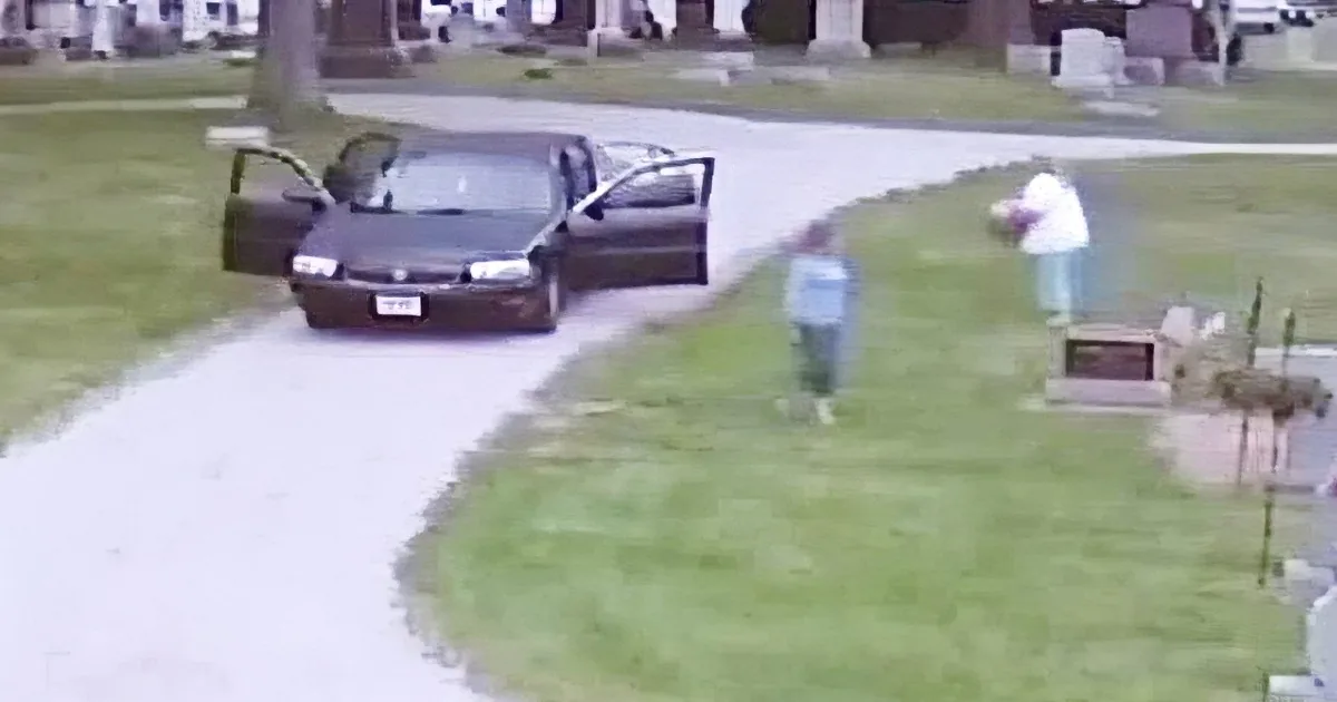 A black car with open doors is parked near a cemetery, with two people walking nearby.