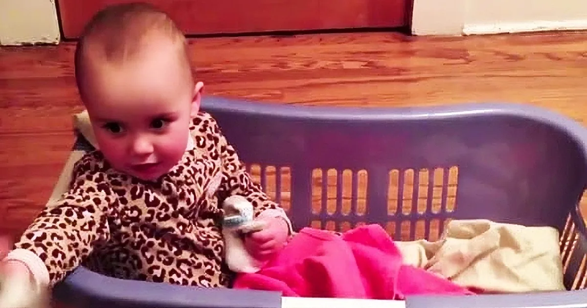 A baby in a leopard print outfit sits in a blue laundry basket with clothes inside.