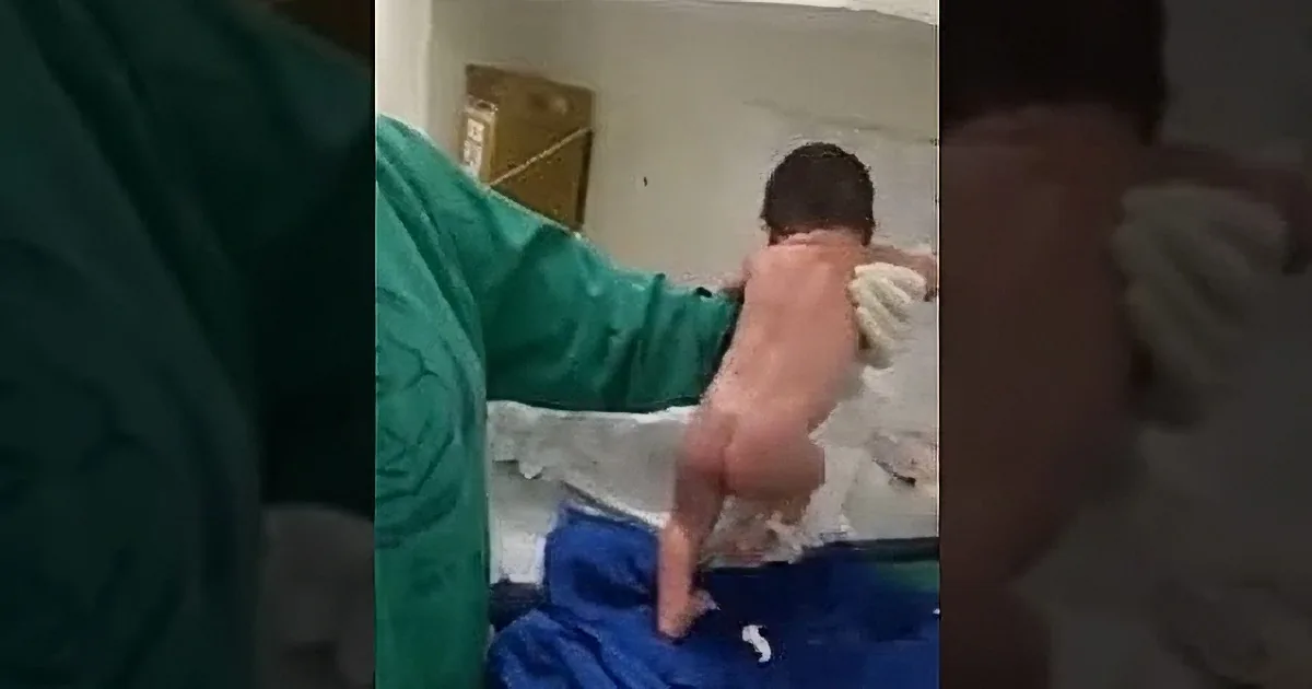 A baby is being held by someone in a green uniform, with a blue blanket visible.