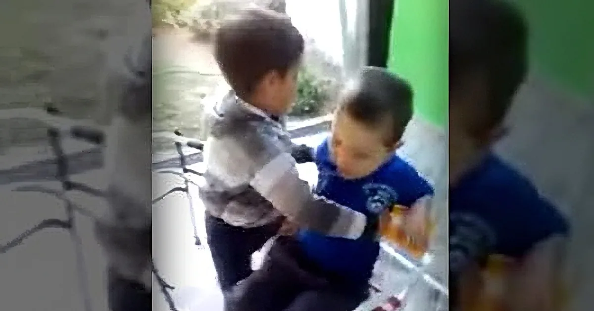 A young boy in a striped shirt sits on a chair, facing another child in a blue shirt who is holding a microphone.