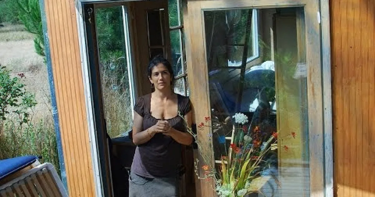 A woman stands in a doorway, hands clasped, surrounded by wooden walls and outdoor scenery.