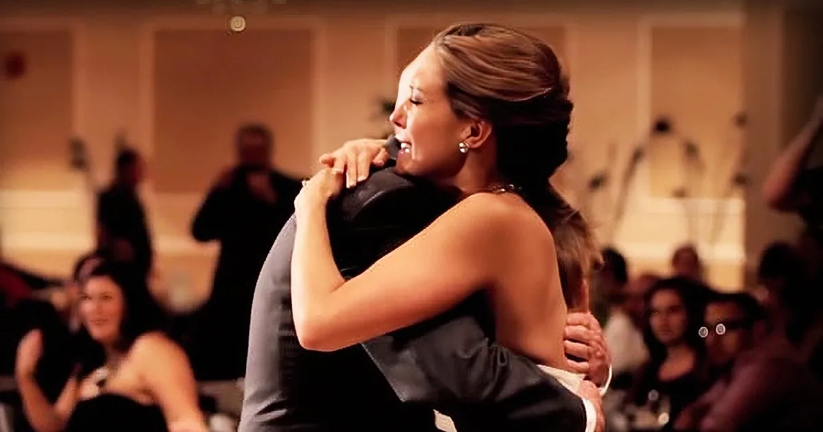 A woman in a black dress hugs a man in a suit, surrounded by an audience in a dimly lit room.