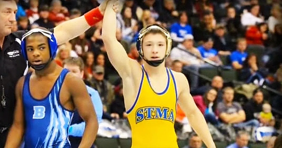 A wrestler in a yellow uniform raises his arm in victory, celebrating a win in a wrestling match.