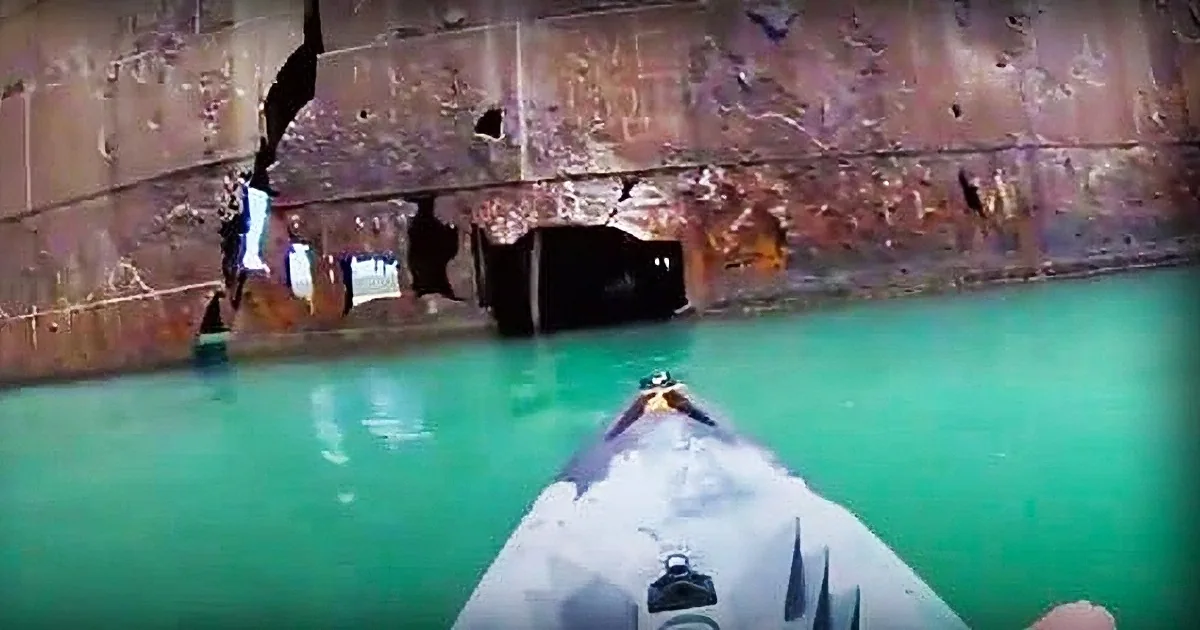A diver in white gear explores a submerged, rusted structure in clear, greenish water.