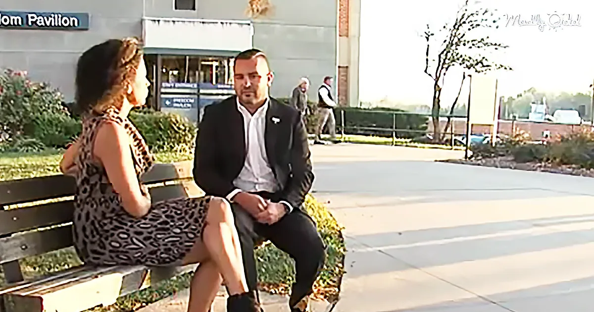 A man and woman sit on a bench, engaged in conversation, with a building and people in the background.
