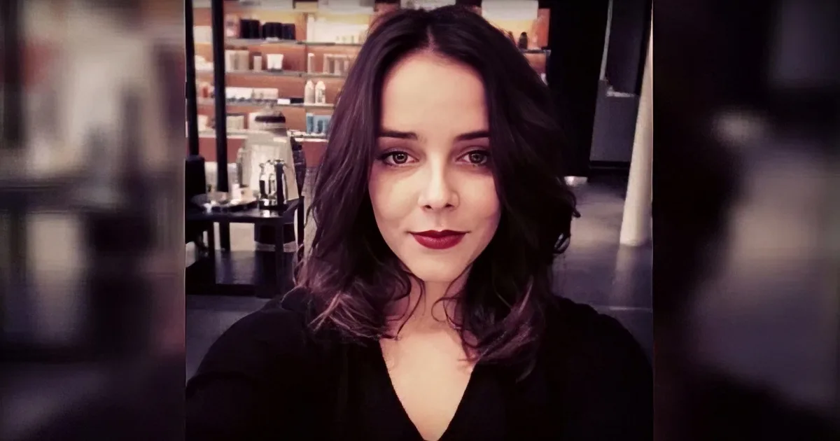 A woman with dark hair and red lipstick, wearing a black top, stands in a blurred indoor setting.