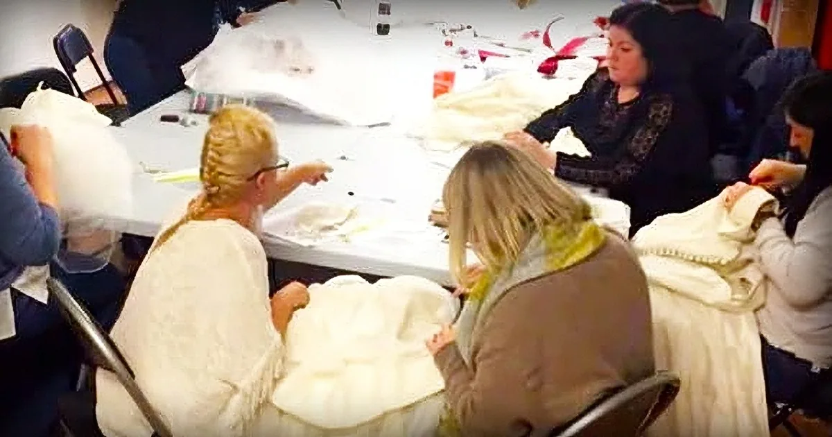 A woman is working on a white garment at a table surrounded by fabric and sewing tools.