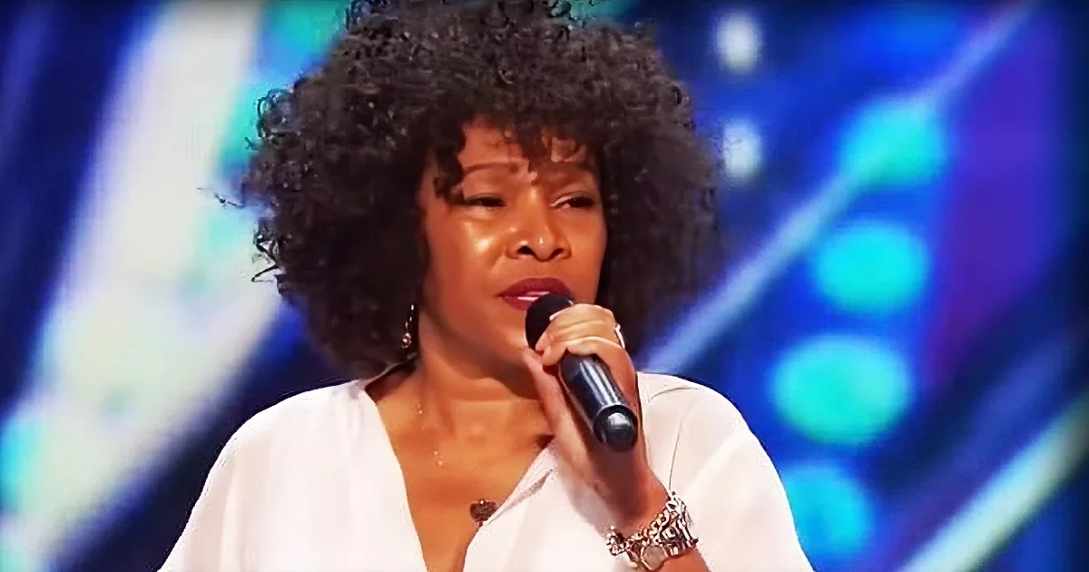 A woman with curly hair, wearing a white top, sings into a microphone against a vibrant blue background.