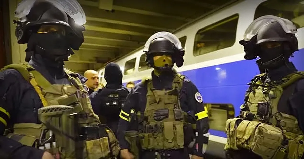Three heavily armed, masked individuals in tactical gear stand in a train station, facing each other.