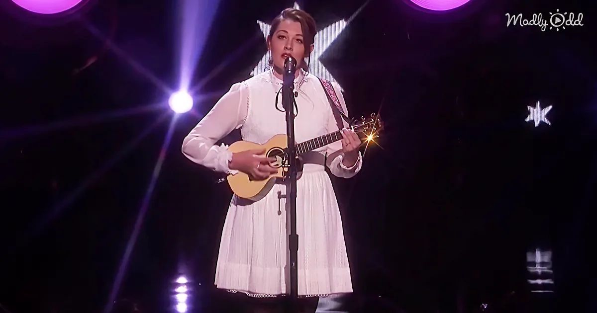A woman in a white dress plays a ukulele on stage with a microphone and purple lights.