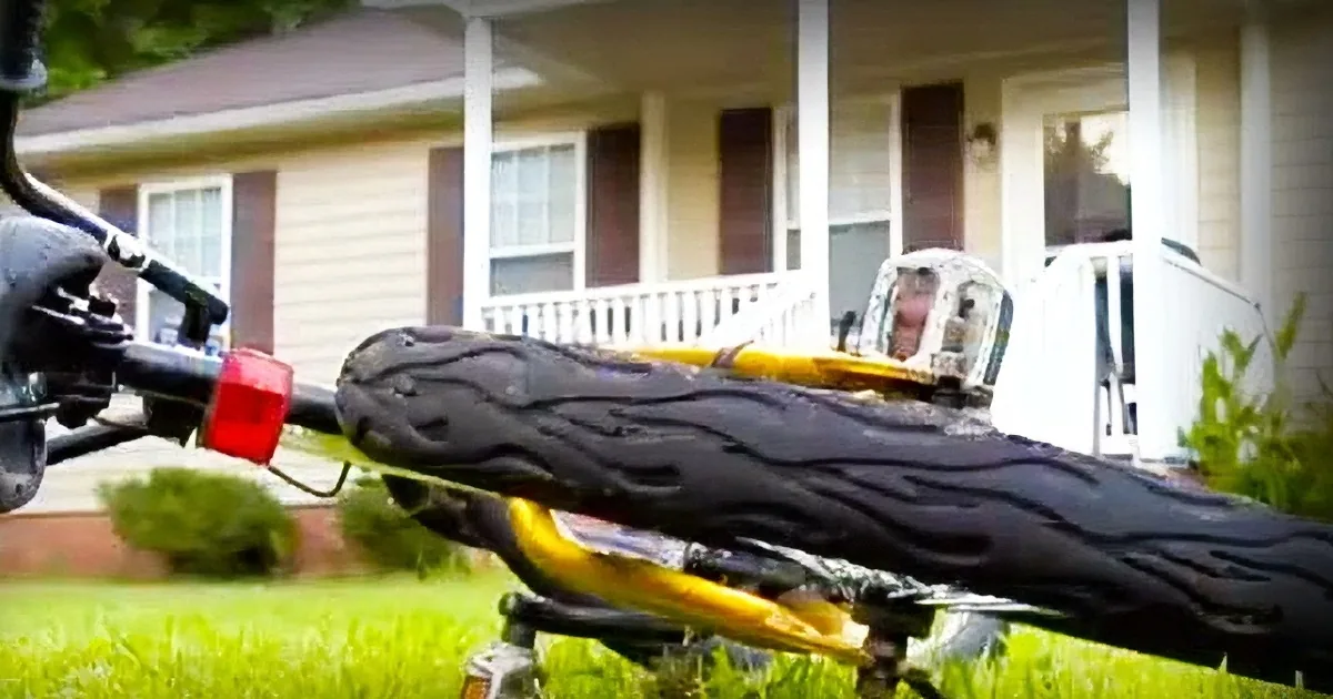 A close-up of a motorcycle's handlebars with a red reflector, set against a suburban backdrop with a house and porch.