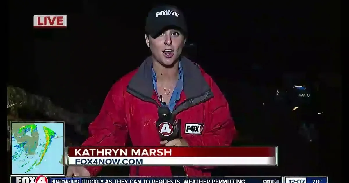 A news reporter in a red jacket and cap is reporting live from a stormy location.