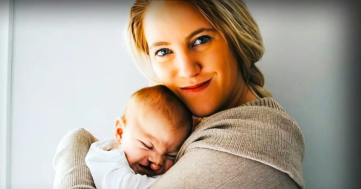 A woman holds a newborn baby, both smiling warmly, in a cozy indoor setting.
