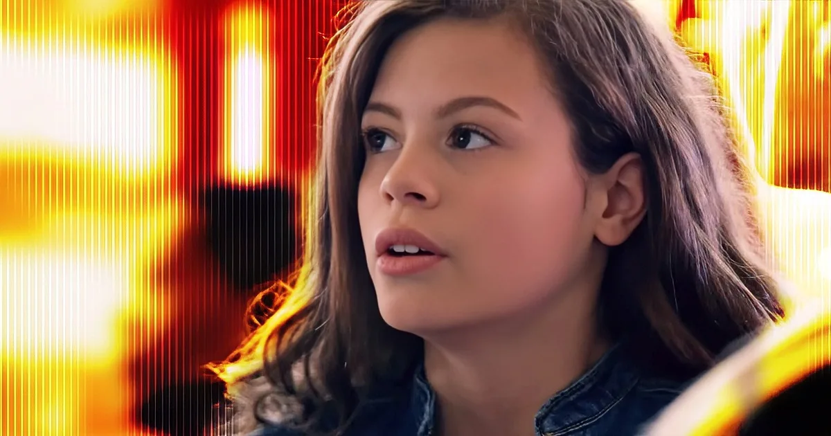A person with long brown hair and a denim jacket, set against a bright, fiery background.