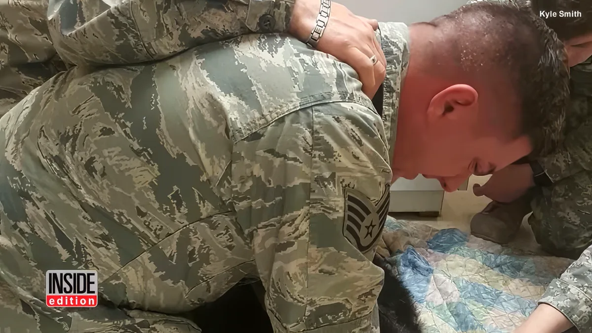 A soldier in camouflage uniform is kneeling, holding another soldier's head, possibly in a moment of support or medical...