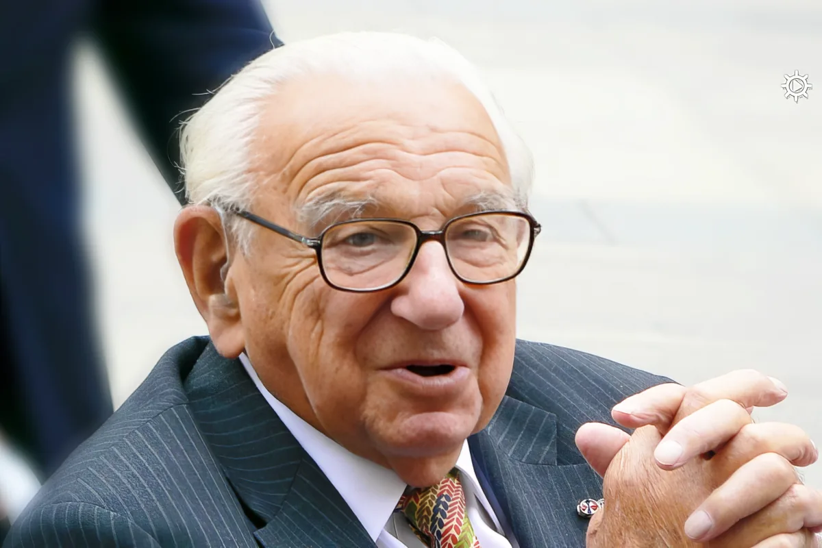 An older man in a suit and tie, wearing glasses, with a hand gesture, set against a blurred background.