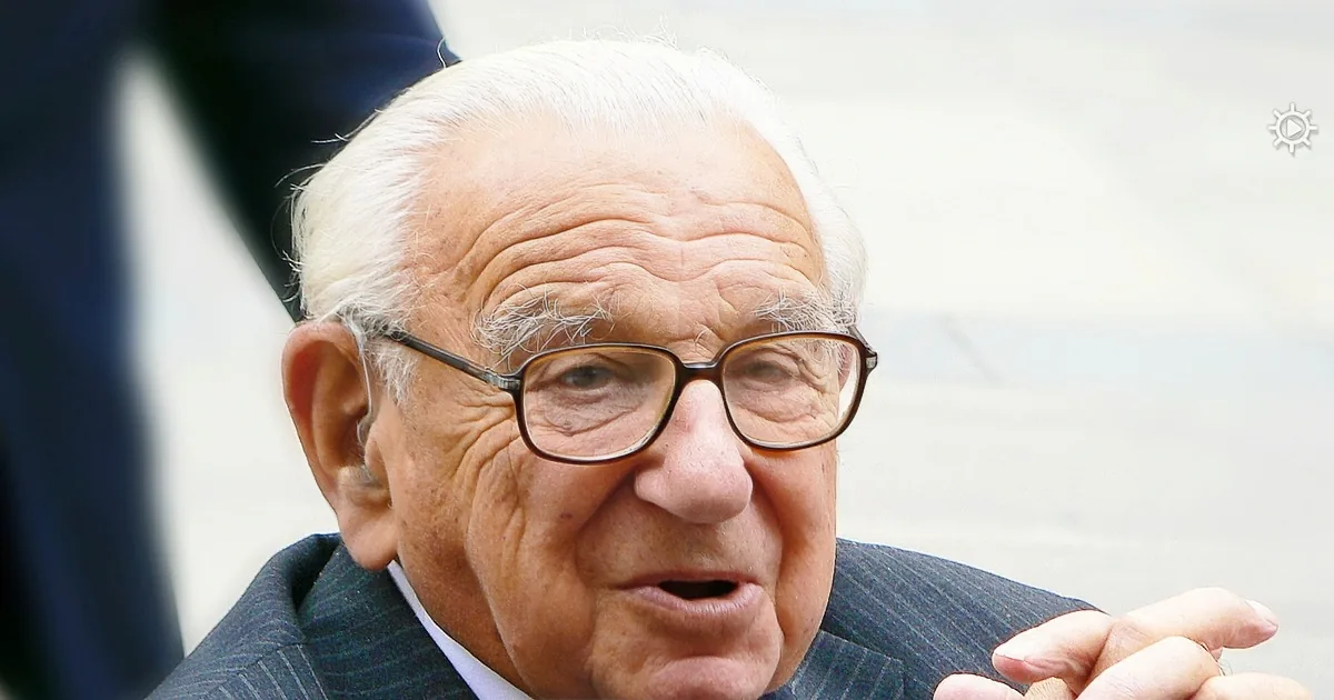 An older man in a suit and tie, wearing glasses, with his hands clasped together.