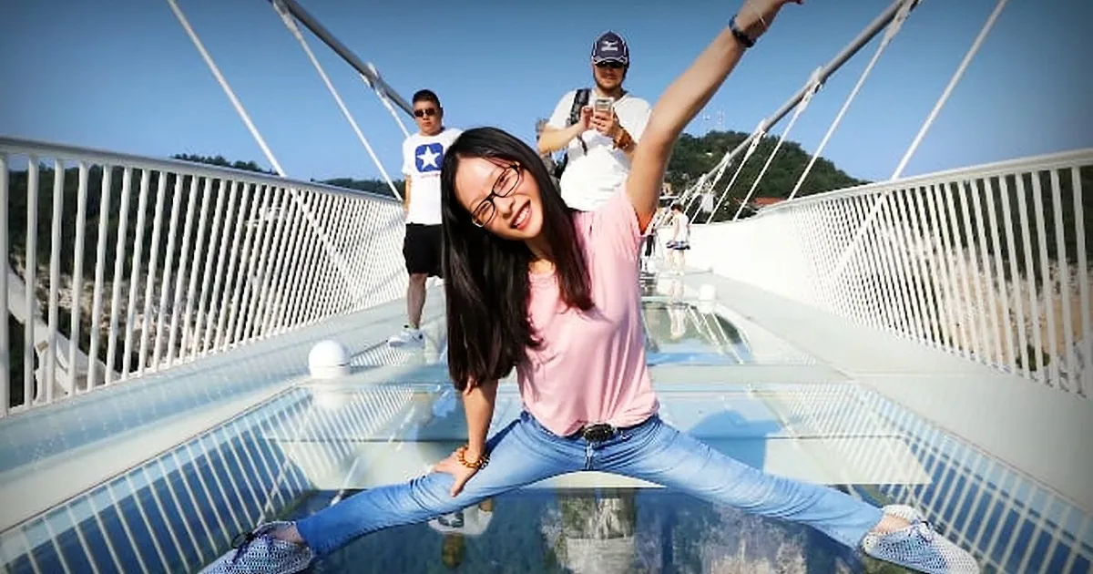 A woman in a pink shirt and jeans strikes a dramatic pose on a glass bridge, with two men in the background.