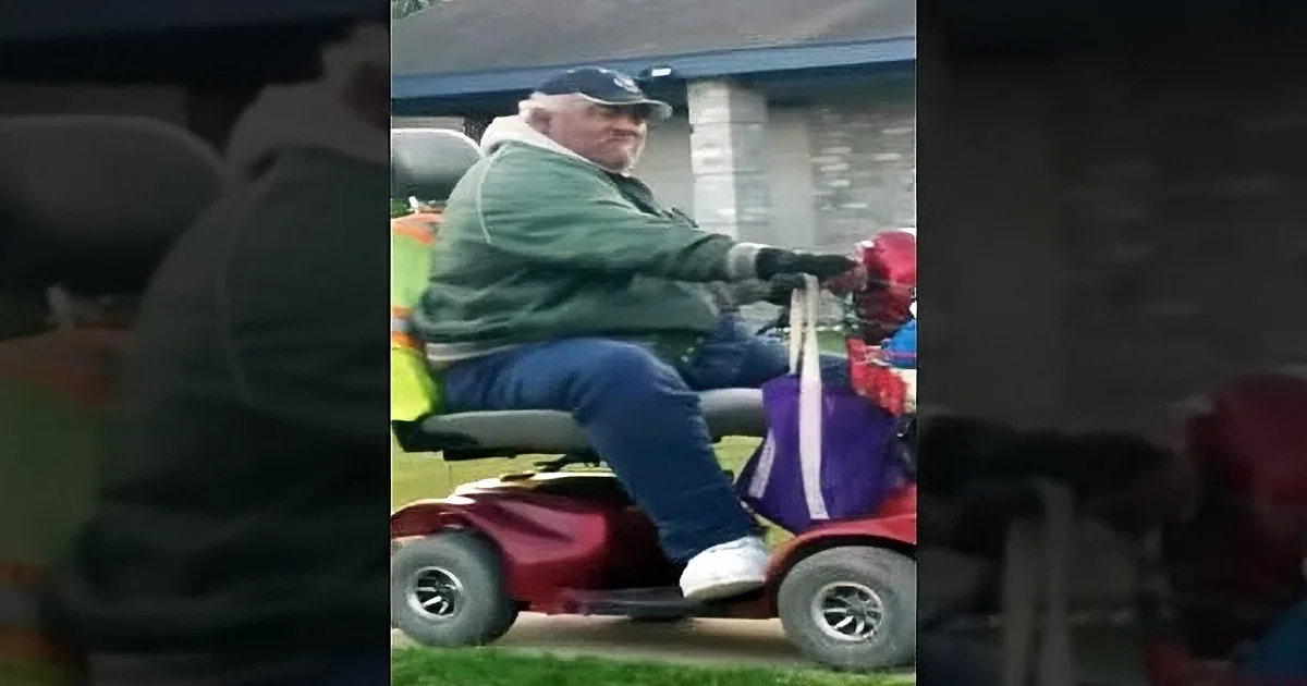 An older man in a green jacket and blue pants rides a red toy car on a grassy area near a house.