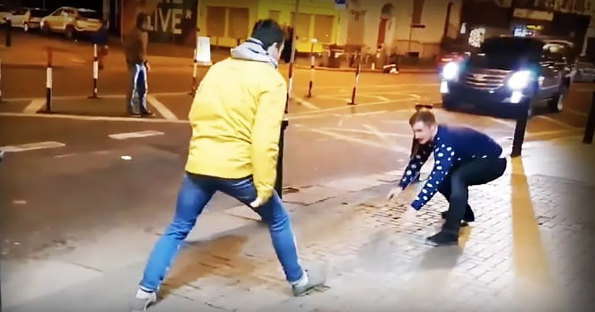 A man in a yellow jacket walks past another man in a blue polka-dot shirt who is crouching on the sidewalk.