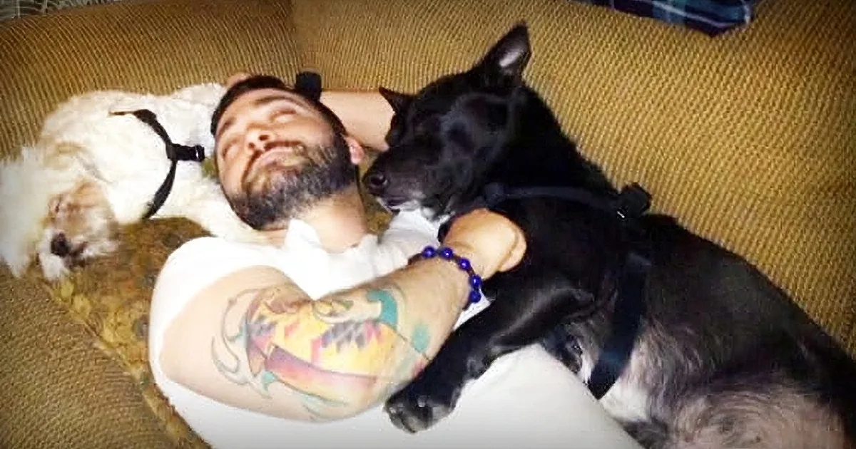 A man with tattoos lies on a couch, cuddling with a black dog and a white dog.