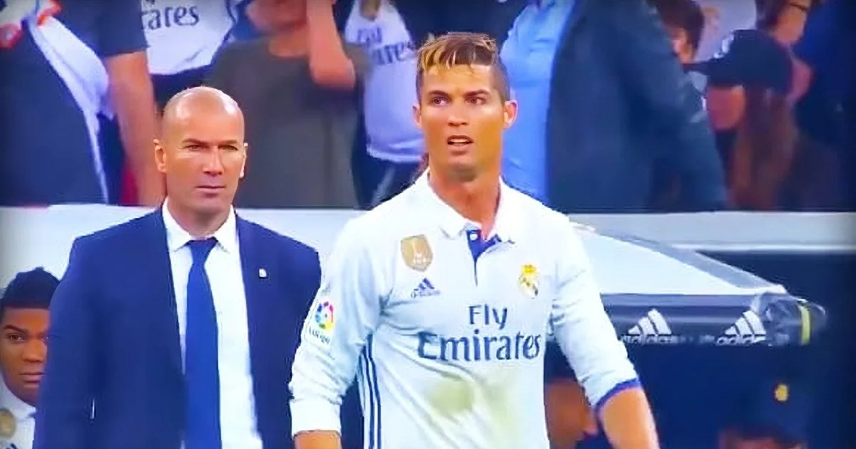 A soccer player in a white jersey with "Emirates" logo is surrounded by other individuals in a stadium setting.