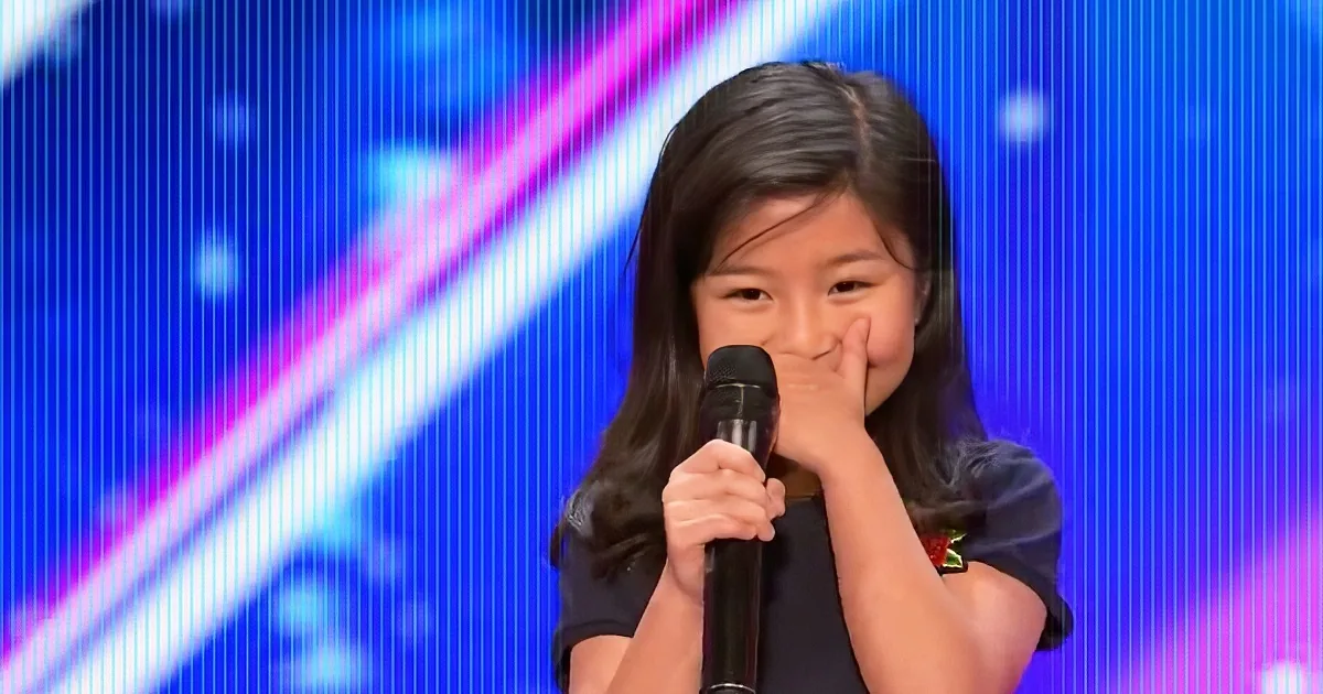 A young girl with long dark hair holds a microphone, covering her mouth with her hand, against a colorful stage backdrop.