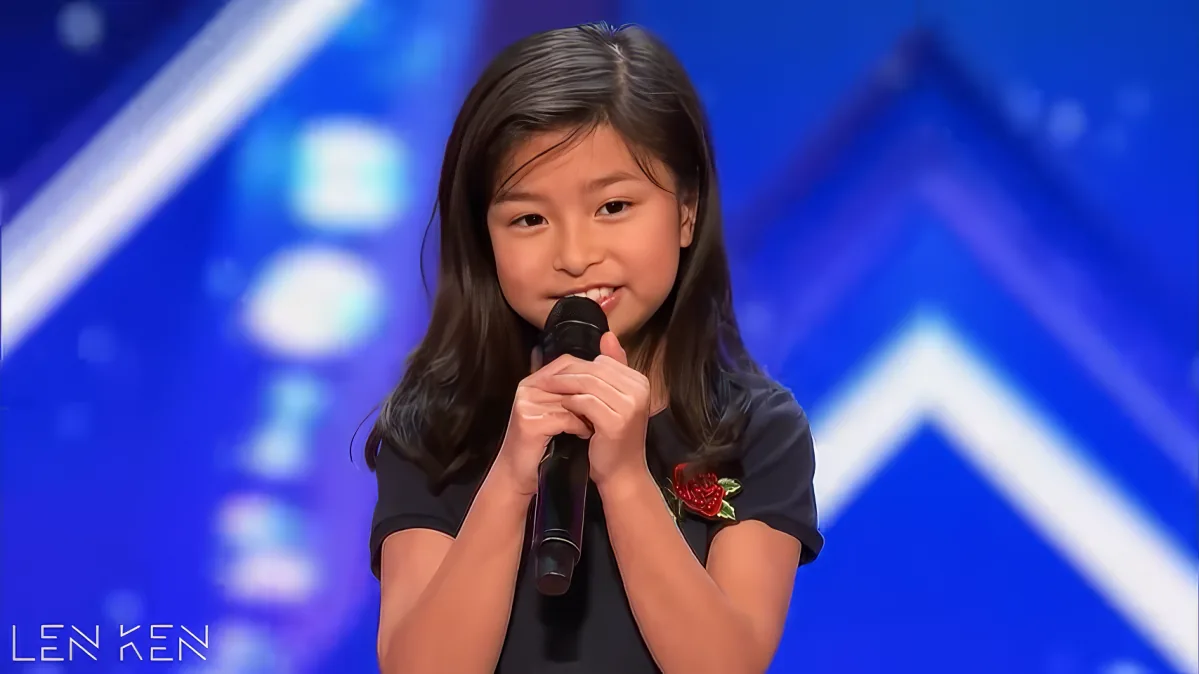 A young girl with long dark hair holds a microphone, wearing a black shirt with a red flower, standing on a stage with a...