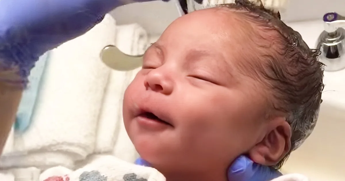A newborn baby with wet hair is being held, with a gloved hand and a sink in the background.