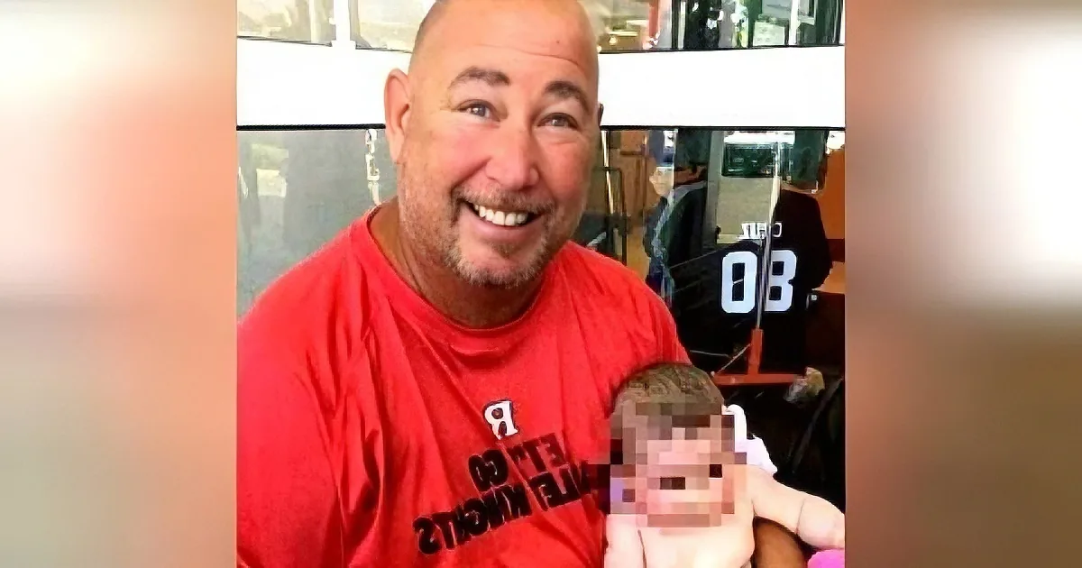 A man in a red shirt smiles while holding a child with blurred face in a public setting.